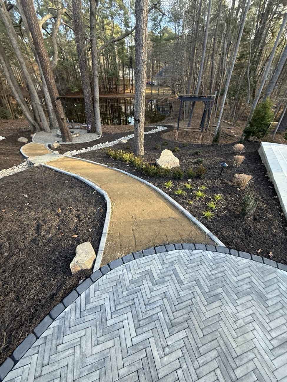 A herringbone paver terrace flowing into a decomposed-granite path that winds through a wooded backyard to a pond.